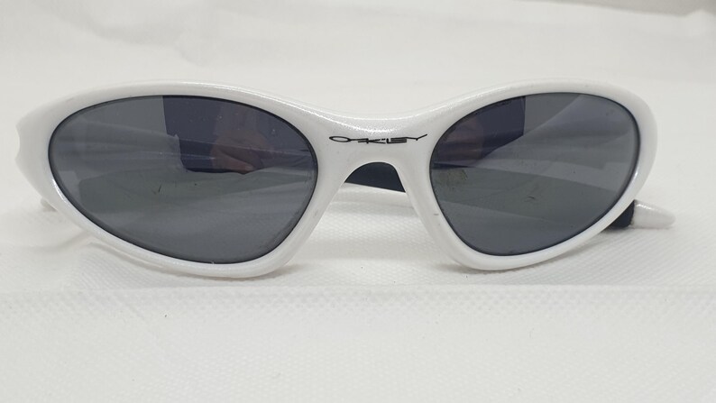 Puede incluir: Gafas de sol blancas de forma ovalada con lentes grises oscuras. Las gafas tienen una montura blanca con el nombre de la marca "Oakley" impreso en negro en el puente. Dise&ntilde;adas para la protecci&oacute;n ocular.