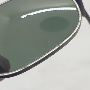 Puede incluir: Primer plano de gafas de sol vintage con montura plateada y lentes verdes. Las gafas de sol est&aacute;n sujetas por una mano, centr&aacute;ndose en los detalles de la montura y las lentes. Las lentes tienen un ligero tinte verde.