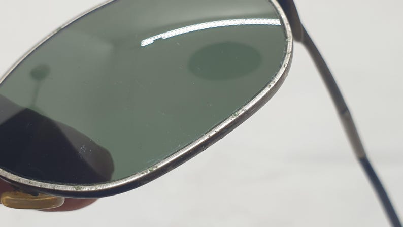 Puede incluir: Primer plano de gafas de sol vintage con montura plateada y lentes verdes. Las gafas de sol est&aacute;n sujetas por una mano, centr&aacute;ndose en los detalles de la montura y las lentes. Las lentes tienen un ligero tinte verde.