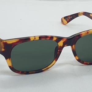 Puede incluir: Gafas de sol carey con lentes verde oscuro. Las monturas tienen un patr&oacute;n cl&aacute;sico marr&oacute;n y amarillo. Las patillas se curvan ligeramente y las lentes son ovaladas. Un peque&ntilde;o logotipo es visible.