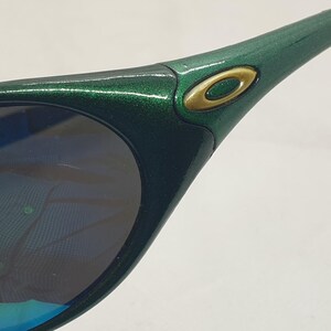 May include: Close-up of green sunglasses with a metallic finish. The sunglasses have a gold-colored logo on the side and dark lenses with a blue and purple reflection. The frame is a dark green color.