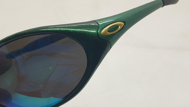 May include: Close-up of green sunglasses with a metallic finish. The sunglasses have a gold-colored logo on the side and dark lenses with a blue and purple reflection. The frame is a dark green color.