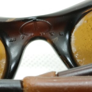 Puede incluir: Primer plano de gafas de sol vintage con montura marr&oacute;n y lentes de color &aacute;mbar. El puente de las gafas presenta texto en relieve y un logotipo. Las lentes muestran algo de desgaste.