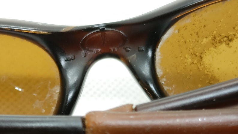 Puede incluir: Primer plano de gafas de sol vintage con montura marr&oacute;n y lentes de color &aacute;mbar. El puente de las gafas presenta texto en relieve y un logotipo. Las lentes muestran algo de desgaste.
