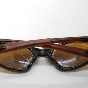 Puede incluir: Gafas de sol marrones y negras con un dise&ntilde;o de montura angular &uacute;nico. Las lentes son de color &aacute;mbar c&aacute;lido y las patillas est&aacute;n plegadas. Las gafas de sol tienen una est&eacute;tica vintage.