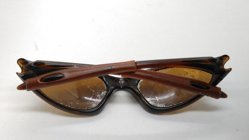 Puede incluir: Gafas de sol marrones y negras con un dise&ntilde;o de montura angular &uacute;nico. Las lentes son de color &aacute;mbar c&aacute;lido y las patillas est&aacute;n plegadas. Las gafas de sol tienen una est&eacute;tica vintage.