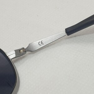 K&ouml;nnte beinhalten: Nahaufnahme einer Sonnenbrille mit silbernem Rahmen und dunklen Gl&auml;sern. Der B&uuml;gel ist schwarz und mit einem Scharnier am Rahmen befestigt. Die Buchstaben "CE" sind auf dem Rahmen aufgedruckt.