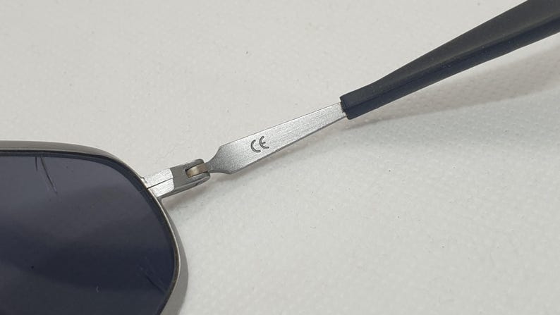 K&ouml;nnte beinhalten: Nahaufnahme einer Sonnenbrille mit silbernem Rahmen und dunklen Gl&auml;sern. Der B&uuml;gel ist schwarz und mit einem Scharnier am Rahmen befestigt. Die Buchstaben "CE" sind auf dem Rahmen aufgedruckt.