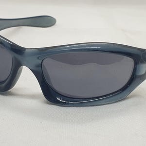 May include: Blue-grey sunglasses with dark lenses. The sunglasses have a curved frame and arms, with a subtle Oakley logo on the temple. The lenses are a dark grey color, and the frame is a solid blue-grey.