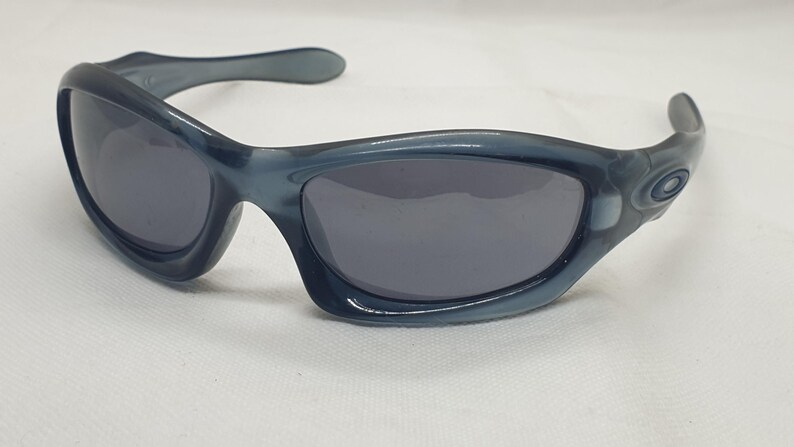 May include: Blue-grey sunglasses with dark lenses. The sunglasses have a curved frame and arms, with a subtle Oakley logo on the temple. The lenses are a dark grey color, and the frame is a solid blue-grey.
