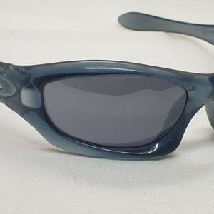 May include: Blue-tinted sunglasses with dark gray lenses. The sunglasses have a curved frame and a small logo on the side. The lenses are a dark gray color, and the frame is a translucent blue.