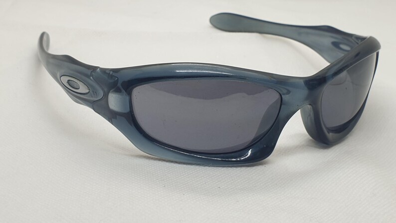 May include: Blue-tinted sunglasses with dark gray lenses. The sunglasses have a curved frame and a small logo on the side. The lenses are a dark gray color, and the frame is a translucent blue.