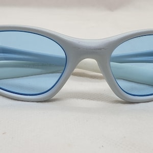 K&ouml;nnte beinhalten: Ovale Sonnenbrille mit hellgrauem Rahmen und blauen Gl&auml;sern. Die Sonnenbrille hat ein schlankes, modernes Design mit einem geschwungenen Steg und B&uuml;geln. Die Gl&auml;ser sind in einem leuchtenden Blau gehalten und bilden einen stilvollen Kontrast zum Rahmen.