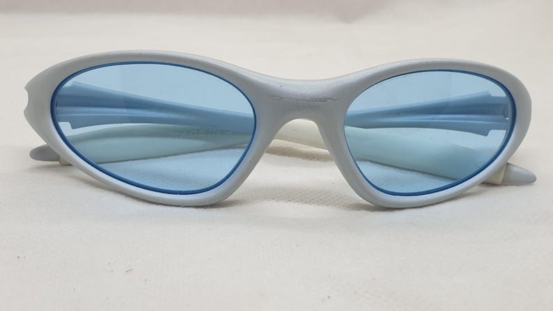 K&ouml;nnte beinhalten: Ovale Sonnenbrille mit hellgrauem Rahmen und blauen Gl&auml;sern. Die Sonnenbrille hat ein schlankes, modernes Design mit einem geschwungenen Steg und B&uuml;geln. Die Gl&auml;ser sind in einem leuchtenden Blau gehalten und bilden einen stilvollen Kontrast zum Rahmen.