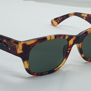 Puede incluir: Gafas de sol de carey con lentes verde oscuro. Las monturas tienen una forma rectangular cl&aacute;sica con un patr&oacute;n marr&oacute;n y amarillo. El logotipo de Ray-Ban es visible en la patilla.