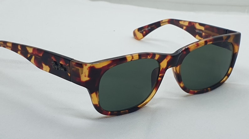 Puede incluir: Gafas de sol de carey con lentes verde oscuro. Las monturas tienen una forma rectangular cl&aacute;sica con un patr&oacute;n marr&oacute;n y amarillo. El logotipo de Ray-Ban es visible en la patilla.