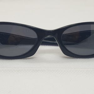 May include: Black cat-eye sunglasses with dark lenses and a glossy finish. The arms of the sunglasses are thin and curve slightly. The sunglasses are set against a white background.