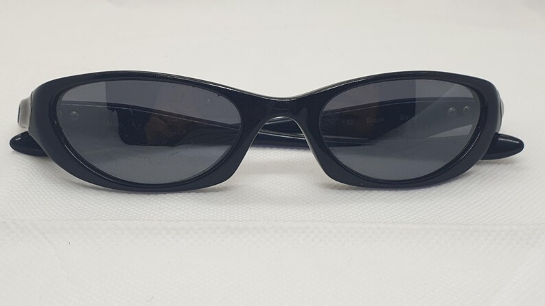 May include: Black cat-eye sunglasses with dark lenses and a glossy finish. The arms of the sunglasses are thin and curve slightly. The sunglasses are set against a white background.