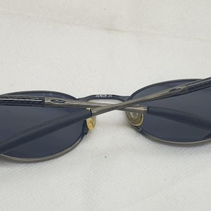 May include: Oval-shaped sunglasses with dark lenses and a dark gray metal frame. The arms are folded across the lenses. The sunglasses are on a white surface.