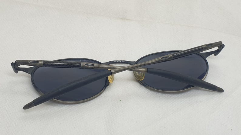 May include: Oval-shaped sunglasses with dark lenses and a dark gray metal frame. The arms are folded across the lenses. The sunglasses are on a white surface.