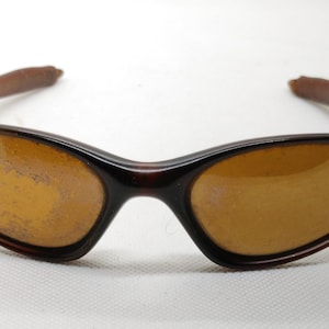 Puede incluir: Gafas de sol marrones con montura curvada y lentes color &aacute;mbar. Las patillas son de un color marr&oacute;n m&aacute;s oscuro. Las gafas de sol est&aacute;n dise&ntilde;adas para proteger los ojos del sol.