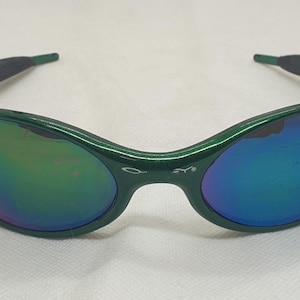 May include: Green-framed sunglasses with iridescent lenses. The lenses reflect a spectrum of colors, including blue, purple, and green. The temples have a dark gray rubberized grip for comfort. The sunglasses are set against a white background.