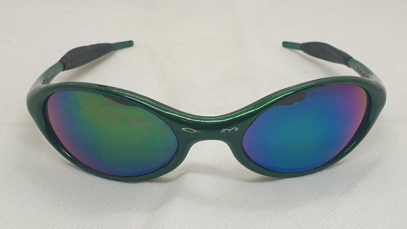 May include: Green-framed sunglasses with iridescent lenses. The lenses reflect a spectrum of colors, including blue, purple, and green. The temples have a dark gray rubberized grip for comfort. The sunglasses are set against a white background.