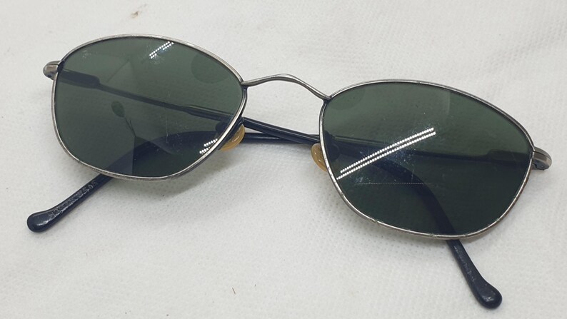 Puede incluir: Gafas de sol vintage con montura de metal plateado y lentes verde oscuro. Las lentes ovaladas est&aacute;n en una montura fina con varillas negras. Un accesorio de gafas cl&aacute;sico.
