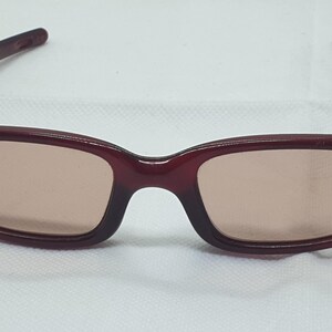 May include: Burgundy sunglasses with rectangular frames and tinted lenses. The glasses have a classic design with a bridge and arms. The lenses are a light brown colour, and the frames are a solid, dark red.