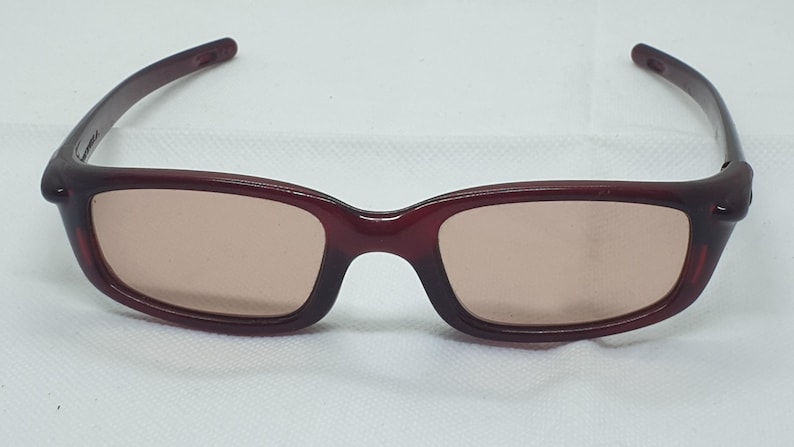May include: Burgundy sunglasses with rectangular frames and tinted lenses. The glasses have a classic design with a bridge and arms. The lenses are a light brown colour, and the frames are a solid, dark red.