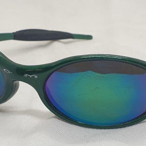 May include: Green sunglasses with iridescent lenses. The oval-shaped frames have a dark green finish, with a black rubberized section on the temples. The lenses reflect a spectrum of colors, including blue, green, and purple.