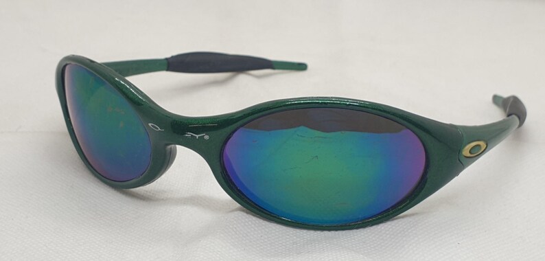 May include: Green sunglasses with iridescent lenses. The oval-shaped frames have a dark green finish, with a black rubberized section on the temples. The lenses reflect a spectrum of colors, including blue, green, and purple.