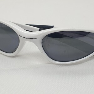 Puede incluir: Gafas de sol blancas con lentes oscuras. Las gafas tienen un dise&ntilde;o elegante y curvado con el nombre de la marca "Oakley" impreso en la montura. Las patillas son negras y las lentes son de color gris oscuro.