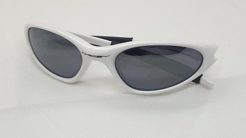Puede incluir: Gafas de sol blancas con lentes oscuras. Las gafas tienen un dise&ntilde;o elegante y curvado con el nombre de la marca "Oakley" impreso en la montura. Las patillas son negras y las lentes son de color gris oscuro.