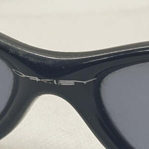 Puede incluir: Primer plano de gafas de sol negras con lentes grises. El nombre de la marca "Oakley" se muestra en letras plateadas en la montura. Las gafas de sol tienen un dise&ntilde;o elegante y moderno.