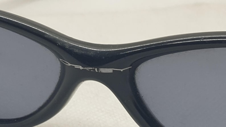 Puede incluir: Primer plano de gafas de sol negras con lentes grises. El nombre de la marca "Oakley" se muestra en letras plateadas en la montura. Las gafas de sol tienen un dise&ntilde;o elegante y moderno.