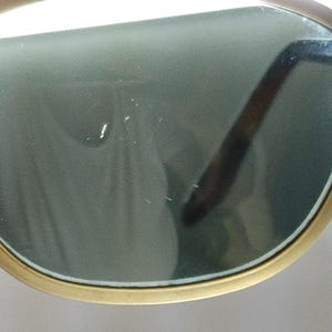 May include: Close-up of gold-rimmed sunglasses with green-tinted lenses. The sunglasses have a classic design with a thin, rectangular frame and dark brown arms. The lenses are slightly reflective, and the overall look is vintage.