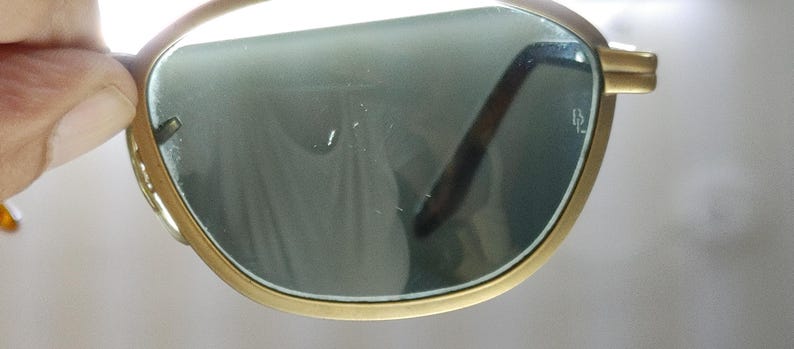 May include: Close-up of gold-rimmed sunglasses with green-tinted lenses. The sunglasses have a classic design with a thin, rectangular frame and dark brown arms. The lenses are slightly reflective, and the overall look is vintage.