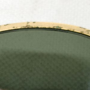 May include: Close-up of vintage sunglasses with a gold-colored frame and green lenses. The frame shows signs of wear, with some discoloration. The text "B&L RAY-" is visible on the temple.