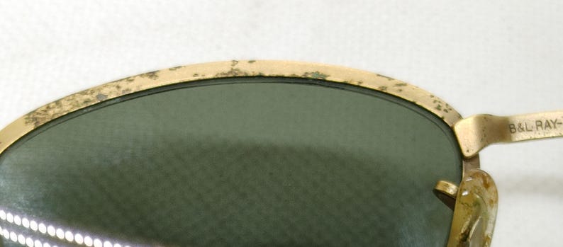 May include: Close-up of vintage sunglasses with a gold-colored frame and green lenses. The frame shows signs of wear, with some discoloration. The text "B&L RAY-" is visible on the temple.