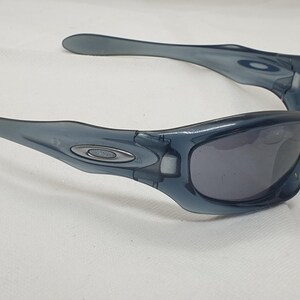 May include: A pair of blue-grey sunglasses with a curved frame and dark lenses. The sunglasses have a sleek, modern design with a small silver emblem on the temples. The arms curve outward for a comfortable fit.