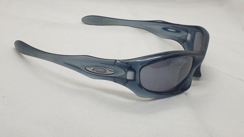 May include: A pair of blue-grey sunglasses with a curved frame and dark lenses. The sunglasses have a sleek, modern design with a small silver emblem on the temples. The arms curve outward for a comfortable fit.