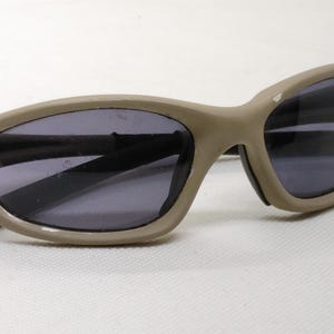 Puede incluir: Gafas de sol color beige con lentes oscuras. Las gafas tienen una forma ovalada y curvada, con una montura gruesa. La montura muestra algo de desgaste. Las lentes son oscuras, proporcionando sombra y protecci&oacute;n solar.