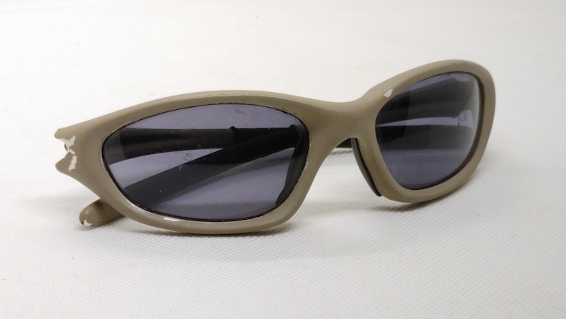 Puede incluir: Gafas de sol color beige con lentes oscuras. Las gafas tienen una forma ovalada y curvada, con una montura gruesa. La montura muestra algo de desgaste. Las lentes son oscuras, proporcionando sombra y protecci&oacute;n solar.