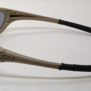 Puede incluir: Gafas de sol color beige con lentes oscuras. Las monturas son de color marr&oacute;n claro con detalles negros en las patillas. El logotipo de Oakley es visible. Un accesorio de moda para la protecci&oacute;n ocular.