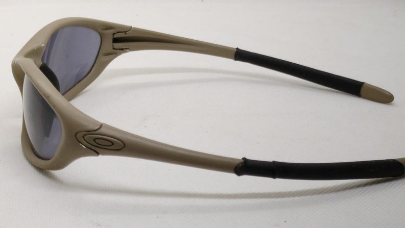 Puede incluir: Gafas de sol color beige con lentes oscuras. Las monturas son de color marr&oacute;n claro con detalles negros en las patillas. El logotipo de Oakley es visible. Un accesorio de moda para la protecci&oacute;n ocular.