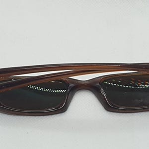 Pode incluir: &Oacute;culos de sol castanhos com arma&ccedil;&atilde;o retangular e lentes escuras. Os &oacute;culos de sol t&ecirc;m um design curvo e est&atilde;o sobre um fundo branco. As hastes dos &oacute;culos s&atilde;o castanho claro.