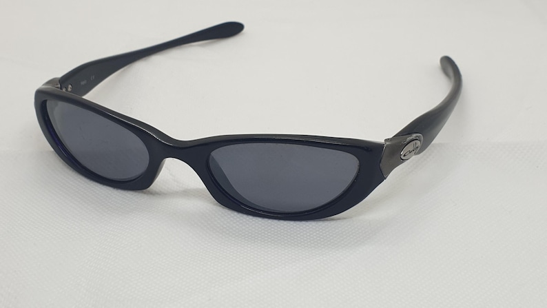 May include: Black sunglasses with a sleek, curved design. The lenses are a dark grey, and the frames are a glossy black. The temples curve gracefully, and the arms are thin. A small, metallic accent is visible on the side of the frame.