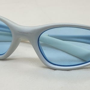 K&ouml;nnte beinhalten: Eine hellgraue Sonnenbrille mit blauen Gl&auml;sern. Die Sonnenbrille hat ein schlankes, geschwungenes Design mit einem d&uuml;nnen Rahmen und einem kleinen Kerbendetail an den &auml;u&szlig;eren Kanten. Die Gl&auml;ser sind hellblau, und die B&uuml;gel sind hellgrau.