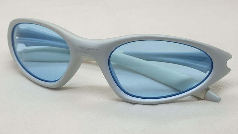 K&ouml;nnte beinhalten: Eine hellgraue Sonnenbrille mit blauen Gl&auml;sern. Die Sonnenbrille hat ein schlankes, geschwungenes Design mit einem d&uuml;nnen Rahmen und einem kleinen Kerbendetail an den &auml;u&szlig;eren Kanten. Die Gl&auml;ser sind hellblau, und die B&uuml;gel sind hellgrau.